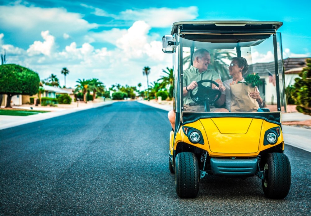 an older husband and wife driving a golf cart on a subdivision street taking their groceries home